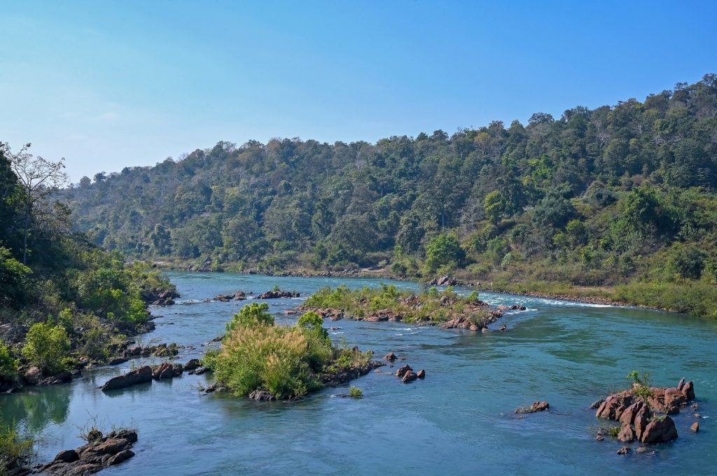 The Hasdeo River in India. Blue river surrounded by green trees with large tree covered rocks in the middle.