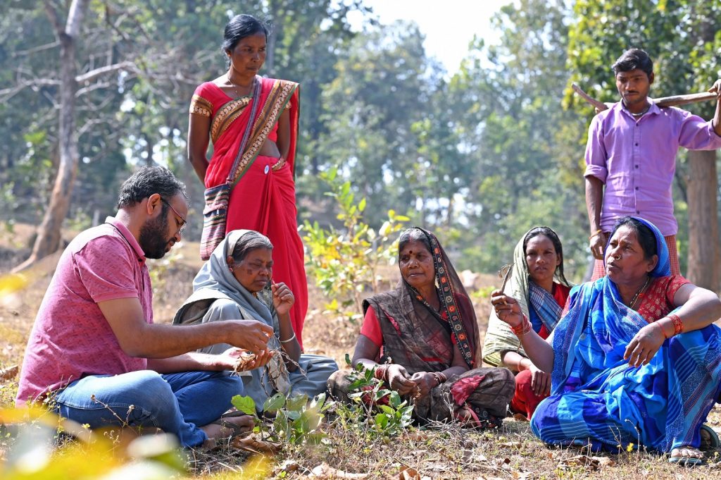 Alok sits with a small group community members wearing brightly colored clothing in the forest.